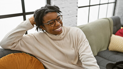 Canvas Print - Cheerful young woman with dreadlocks wearing glasses enjoying time in a cozy living room on a sofa.