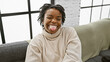 A joyful african american woman with dreadlocks wearing a sweater poses playfully on an indoor sofa.