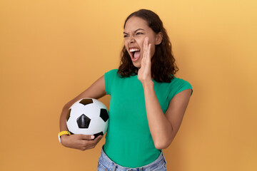 Wall Mural - Young hispanic woman holding ball shouting and screaming loud to side with hand on mouth. communication concept.