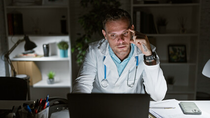 Sticker - Hispanic man in lab coat pondering at night in a medical office with laptop and stethoscope