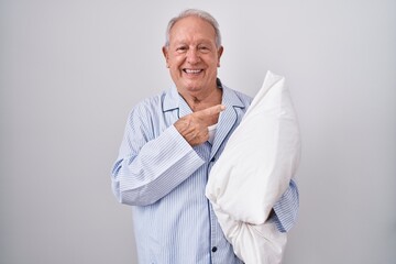 Canvas Print - Senior man with grey hair wearing pijama hugging pillow cheerful with a smile on face pointing with hand and finger up to the side with happy and natural expression