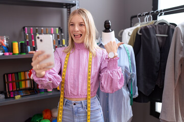 Canvas Print - Young caucasian woman dressmaker designer on video call with smartphone pointing thumb up to the side smiling happy with open mouth