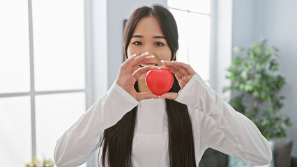 Canvas Print - A smiling asian woman holds a red heart, symbolizing love, in a well-lit, modern living room with minimalist decor.