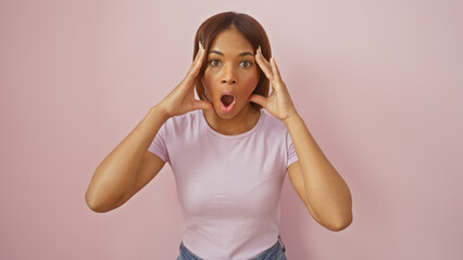 Wall Mural - A surprised african american woman in casual clothing against a pink background expressing amazement with hands on cheeks.