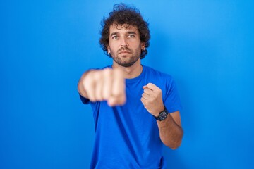 Poster - Hispanic young man standing over blue background punching fist to fight, aggressive and angry attack, threat and violence