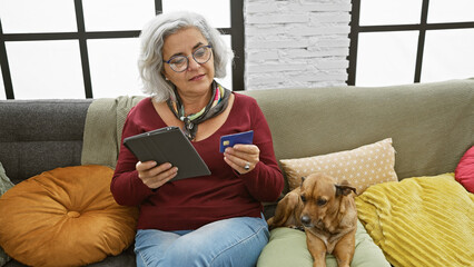 Poster - Mature woman with glasses using tablet and holding credit card, dog beside her, in a cozy living room interior.