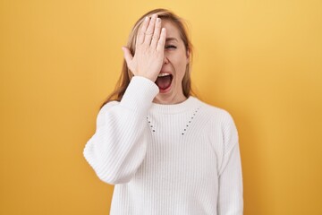 Poster - Young caucasian woman wearing white sweater over yellow background covering one eye with hand, confident smile on face and surprise emotion.