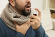 Young man with scarf using throat spray indoors, closeup