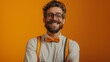 A stylish bearded man wearing glasses, a bow tie, and suspenders smiling confidently on an orange background.