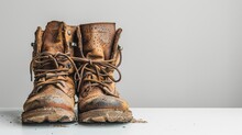A Pair Of Heavily Used Brown Leather Work Boots Covered In Mud, Standing On A White Surface With A Grey Background.