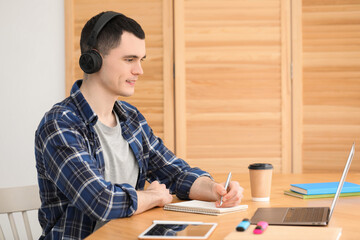 Wall Mural - E-learning. Young man taking notes during online lesson at table indoors.