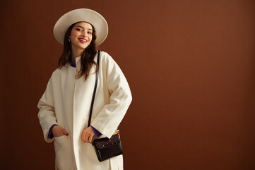 Fashionable happy smiling  beautiful brunette woman wearing trendy hat, white coat, stylish leather shoulder bag, posing on brown background. Studio fashion portrait. Copy, empty, blank space for text