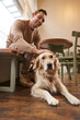 Vertical portrait of smiling handsome man with his dog, drinks coffee in pet-friendly cafe in city, touches golden retriever