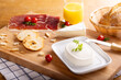 Cream Cheese spread on a black tray with prosciutto, cheese, butter, bread toasts and bread. A glass of orange juice is also served on the table.
