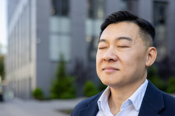 Close-up photo of a young Asian male office worker standing outside, thoughtfully closing his eyes and resting