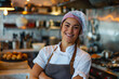Smiling young female chef in apron and chef's hat stands with arms crossed in restaurant kitchen