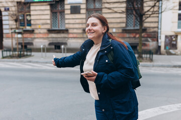 Wall Mural - Stylish 30s woman with backpack holding smart phone and catching a taxi on the city street. Urban lifestyle concept.