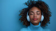 Woman with curly hair enjoying music on headphones, eyes closed, serene against a blue background.
