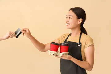 Sticker - Female Asian barista with cups of coffee taking credit card on beige background