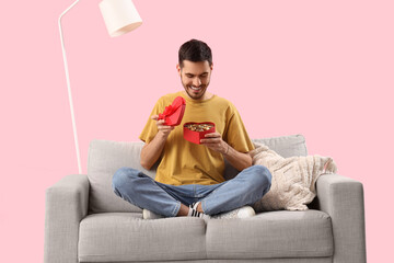 Wall Mural - Young man with box of heart-shaped chocolate candies sitting on sofa against pink background