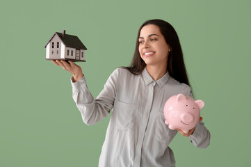 Wall Mural - Beautiful young happy woman with piggy bank and house model on green background. Mortgage concept