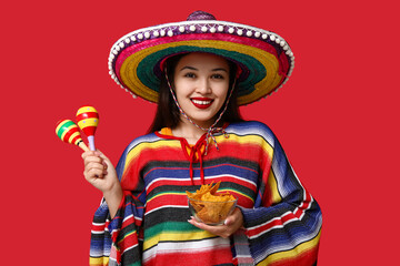 Wall Mural - Portrait of young woman with colorful poncho, sombrero, maracas and tortilla chips on red background. National Tortilla Chip Day celebration