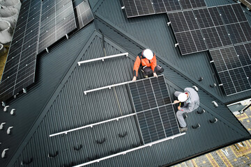 Wall Mural - Mounters building photovoltaic solar module station on roof of house. Men electricians in helmets installing solar panel system outdoors. Concept of alternative and renewable energy. Aerial view.