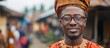 A close-up shot of an Igbo businessman, wearing a turban and glasses, in a village setting. He is standing outdoors, showcasing traditional attire and a sense of cultural identity.