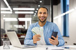 © Tetiana - Portrait of a happy young hispanic man sitting in the office at the table, holding cash money and a phone, smiling at the camera