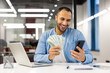 © Tetiana - Smiling hispanic young man sitting in a formal office at a table, holding cash money banknotes in his hands, using a mobile phone