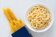 Cooked spaghetti in a white ceramic bowl next to uncooked spaghetti in a blue box on white painted wood.