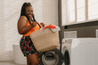 © gstockstudio - Happy plus size African woman carrying basket with clothes while standing near the washing machine in bathroom