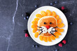 Cute child theme breakfast pancake in the shape of a lion face. Above view table scene on a dark stone background.