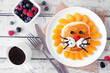 Cute child theme breakfast pancake in the shape of a lion face. Overhead view table scene on a white wood background.