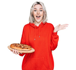 Sticker - Young blonde girl holding italian pizza celebrating victory with happy smile and winner expression with raised hands