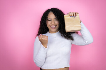 Wall Mural - African american woman wearing casual sweater over pink background holding a paper bag very happy and excited making winner gesture with raised arms, smiling and screaming for success