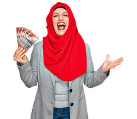 Poster - Beautiful hispanic woman wearing islamic hijab holding australian dollars banknotes celebrating victory with happy smile and winner expression with raised hands