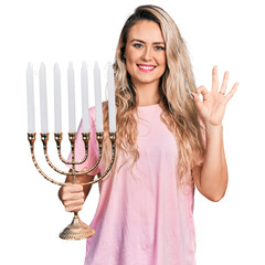 Poster - Young blonde woman holding menorah hanukkah jewish candle doing ok sign with fingers, smiling friendly gesturing excellent symbol