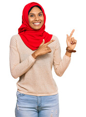 Wall Mural - Young african american woman wearing traditional islamic hijab scarf smiling and looking at the camera pointing with two hands and fingers to the side.