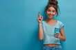 Portrait of beautiful young woman in casual smiling and pointing finger to side while holding smartphone