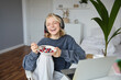 Portrait of happy young blond woman, sitting in a room, watching movie on laptop and eating healthy breakfast, drinking tea, resting on weekend
