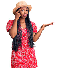 Sticker - Young african american woman wearing summer hat confused and annoyed with open palm showing copy space and pointing finger to forehead. think about it.