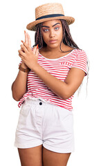 Wall Mural - Young african american woman with braids wearing summer hat holding symbolic gun with hand gesture, playing killing shooting weapons, angry face