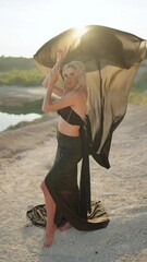 Wall Mural - Young blonde woman performs belly dance at sunset on shore of lake. Woman dressed in black and with jewelry on her head stands barefoot by water. Ethnic beauty concept, sunset, model