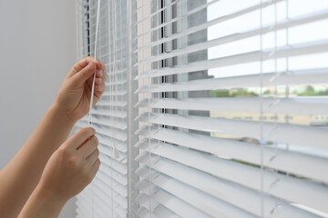 Woman opening horizontal blinds on window indoors, closeup