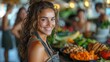 A happy woman is smiling by a table full of natural foods like fruits and leaf vegetables. She seems excited about cooking and creating new recipes with these fresh ingredients