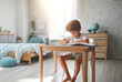 Kind Junge sitzt entspannt in hellem Kinder Jugend Zimmer barfuß am Schreibtisch schreibt lernt Hausaufgaben Schule Bildung zuhause allein spielend lernen glücklich und zufrieden flow motiviert 