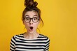 Young student girl very surprised by something. Beautiful nerdy young woman in striped top and glasses isolated on yellow background looking at camera with funny, shocked, astonished face expression