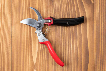 Wall Mural - Steel gardening secateurs, scissors tool with red and black grip for pruned of plants and flowers garden work, on a wooden table background. Open state. Top view.