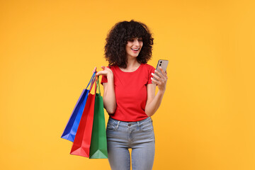 Wall Mural - Happy young woman with shopping bags and modern smartphone on yellow background